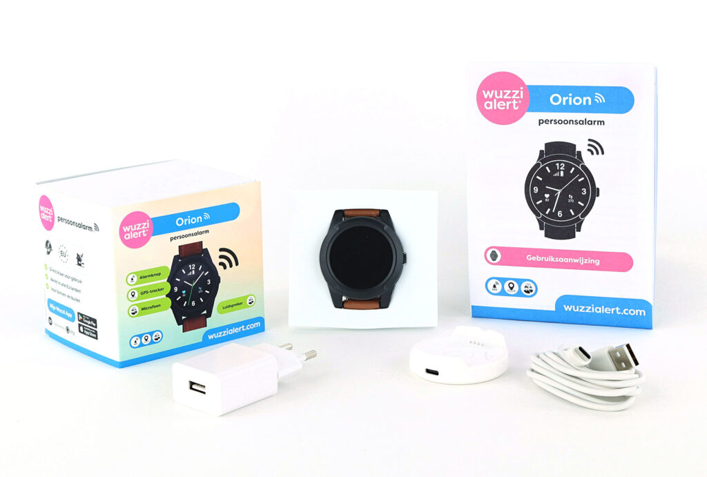unboxing van de verpakking van de Wuzzi Alert Orion alarmhorloge. Waarbij het alarmhorloge in het midden zichtbaar is, omringd door de verpakking, de gebruiksaanwijzing en bijbehorende accessoires zoals het oplaadstation, de oplaadkabel en de witte stekker.